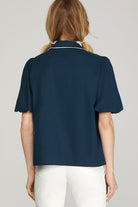 A woman shown from the back wearing a navy blue short-sleeve collared blouse with thin white piping trim and white pants, against a light gray studio background.