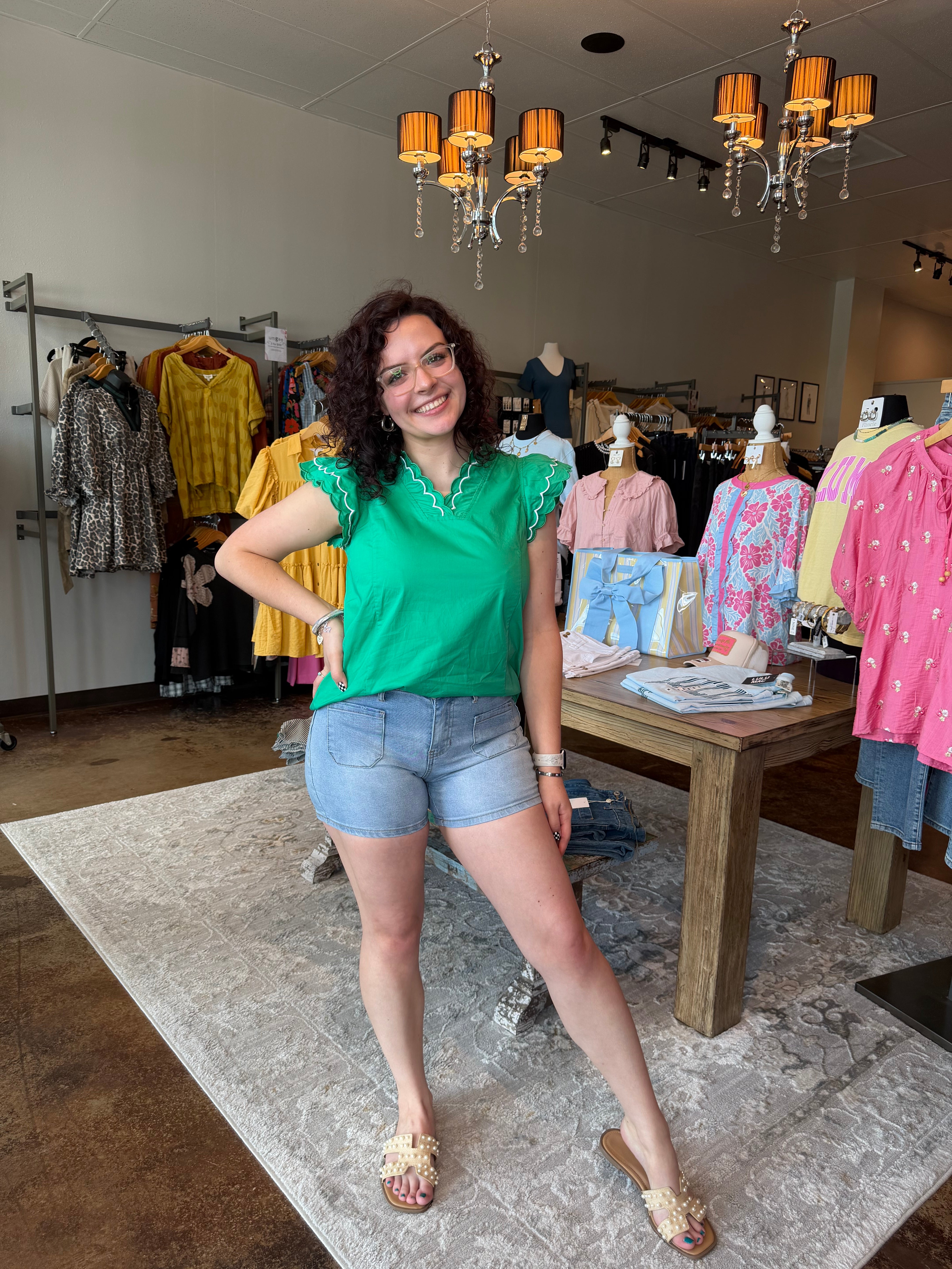 Woman in a green shirt and denim shorts standing in a store with clothing racks and displays.