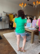 Woman in a green shirt and shorts standing in a clothing store with racks of clothes and a chandelier.