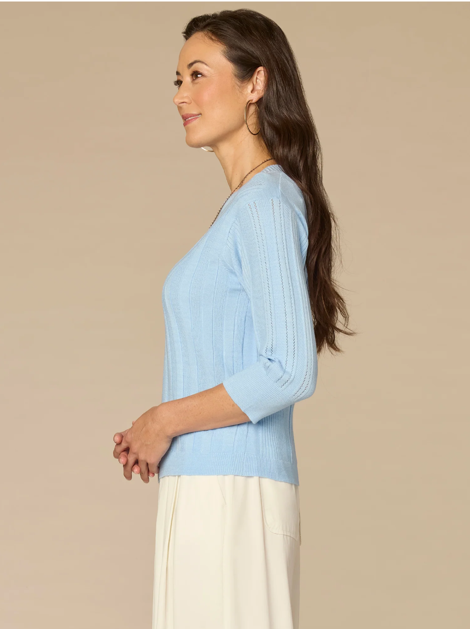 Woman wearing a light blue sweater and white pants against a beige background