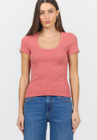 A woman modeling a short-sleeve scoop-neck ribbed knit top in dusty rose pink, paired with blue jeans, against a light gray studio background.