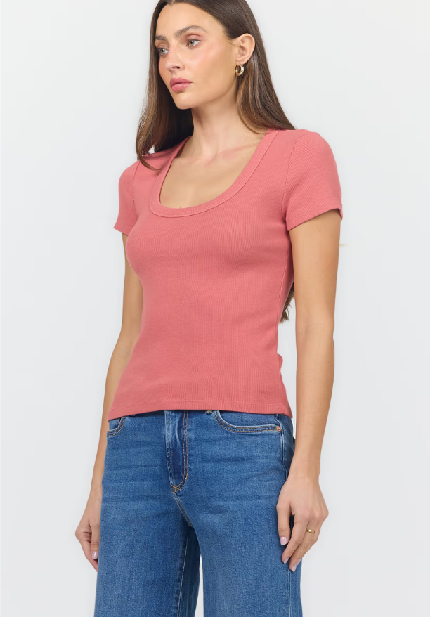 A woman wearing a fitted short-sleeve scoop-neck ribbed knit top in dusty rose pink, paired with blue jeans, against a plain light gray studio background.