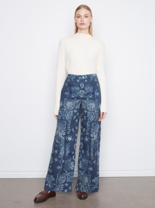 A model stands against a white wall wearing a cream ribbed mock-neck top and high-waisted, wide-leg trousers with a blue and white paisley pattern. She is wearing brown leather loafers.