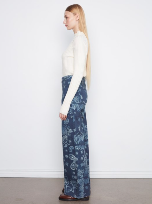 A woman stands in profile wearing a cream ribbed mock-neck long-sleeve top and high-waisted, wide-leg navy blue pants with a light blue paisley pattern, set against a plain white wall and light gray floor.