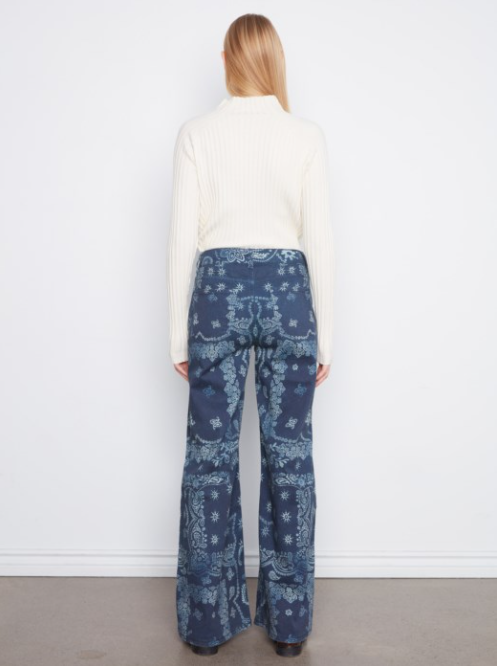 A woman viewed from behind wearing a cream ribbed mock-neck sweater and blue patterned wide-leg pants, standing against a plain white wall.