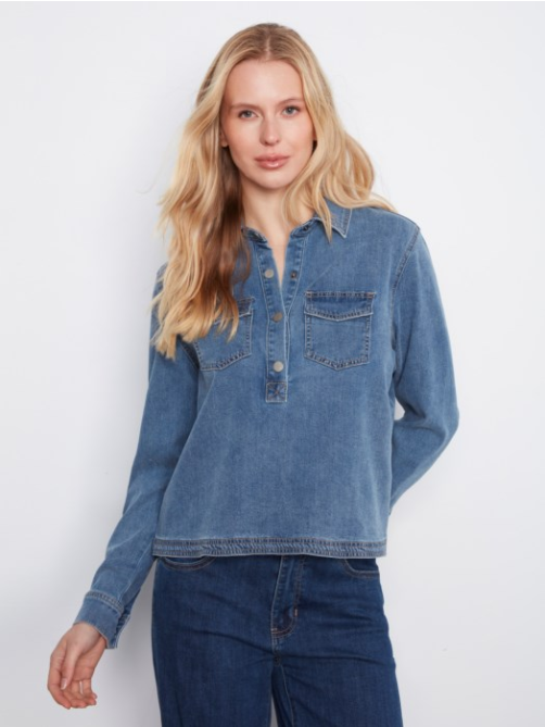 A blonde woman wearing a cropped, long-sleeve blue denim shirt-jacket with a buttoned henley-style placket and a chest pocket, paired with dark blue jeans, against a plain white studio background.