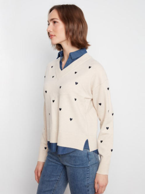 A woman wearing a cream-colored knit sweater with small black heart details, layered over a blue collared shirt and paired with blue jeans, shown from a side profile against a plain white background.