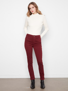 A woman modeling a cream mock-neck long-sleeve top and dark red slim-fit jeans with black ankle boots, standing against a white wall.