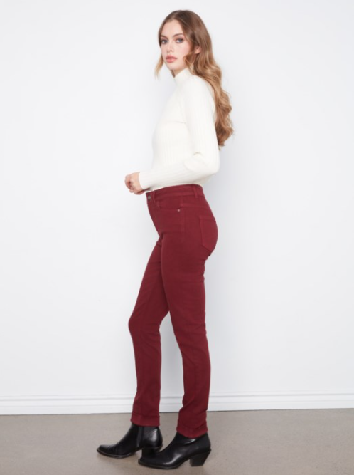 A woman modeling high-waisted, ankle-length burgundy corduroy pants, paired with a white mock-neck top and black ankle boots, standing against a white wall.