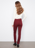A woman with long wavy light brown hair is shown from behind wearing high-waisted, dark red skinny jeans, a cream long-sleeve top, and black ankle boots against a white wall.