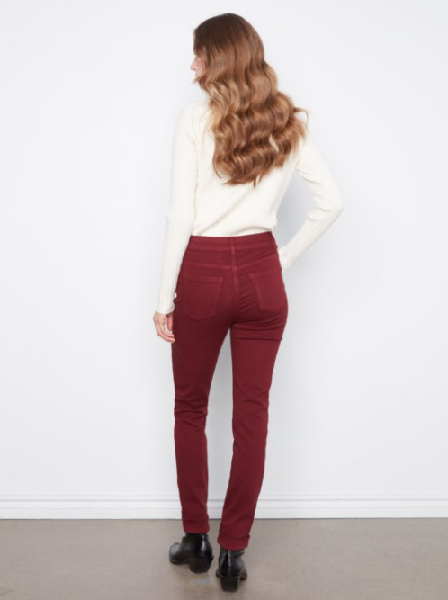 A woman with long wavy light brown hair is shown from behind wearing high-waisted, dark red skinny jeans, a cream long-sleeve top, and black ankle boots against a white wall.