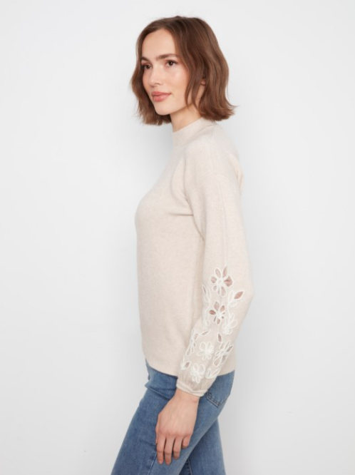 A woman wearing a light beige mock-neck knit sweater with white floral lace embroidery on the sleeve, paired with blue jeans, photographed against a plain white background.