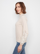 A woman wearing a light beige mock-neck knit sweater with white floral lace embroidery on the sleeve, paired with blue jeans, photographed against a plain white background.