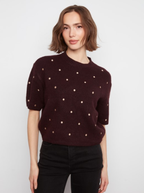 A woman wearing a short-sleeve crewneck knit sweater in deep burgundy with small beige polka dots, paired with black pants, against a light gray studio background.