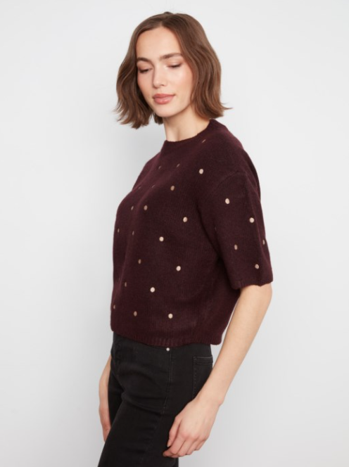A woman wearing a short-sleeve crewneck knit sweater in deep burgundy with small metallic gold polka dots, paired with black jeans, against a plain light gray background.