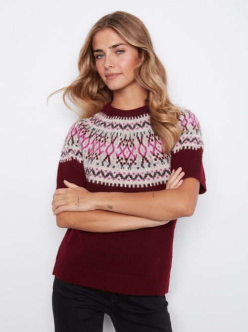 A woman wearing a short-sleeve knit sweater with a burgundy body and a pink, white, and dark brown geometric Fair Isle pattern across the chest and shoulders, paired with black pants, against a plain white background.