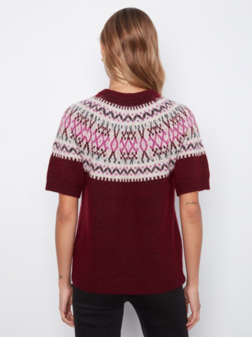 A woman viewed from the back wearing a short-sleeve knit sweater with a deep burgundy body and a colorful Fair Isle yoke pattern in cream, pink, and dark brown, paired with black pants against a plain white background.
