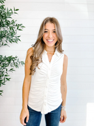 Woman wearing a white sleeveless top and blue jeans standing against a white wall with a plant to the left.
