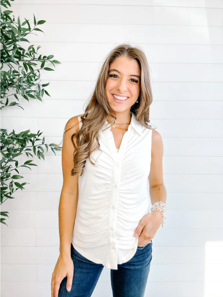 Woman wearing a white sleeveless top and blue jeans against a white brick wall with greenery.