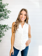 Woman wearing a white sleeveless top and blue jeans against a white brick wall with greenery.