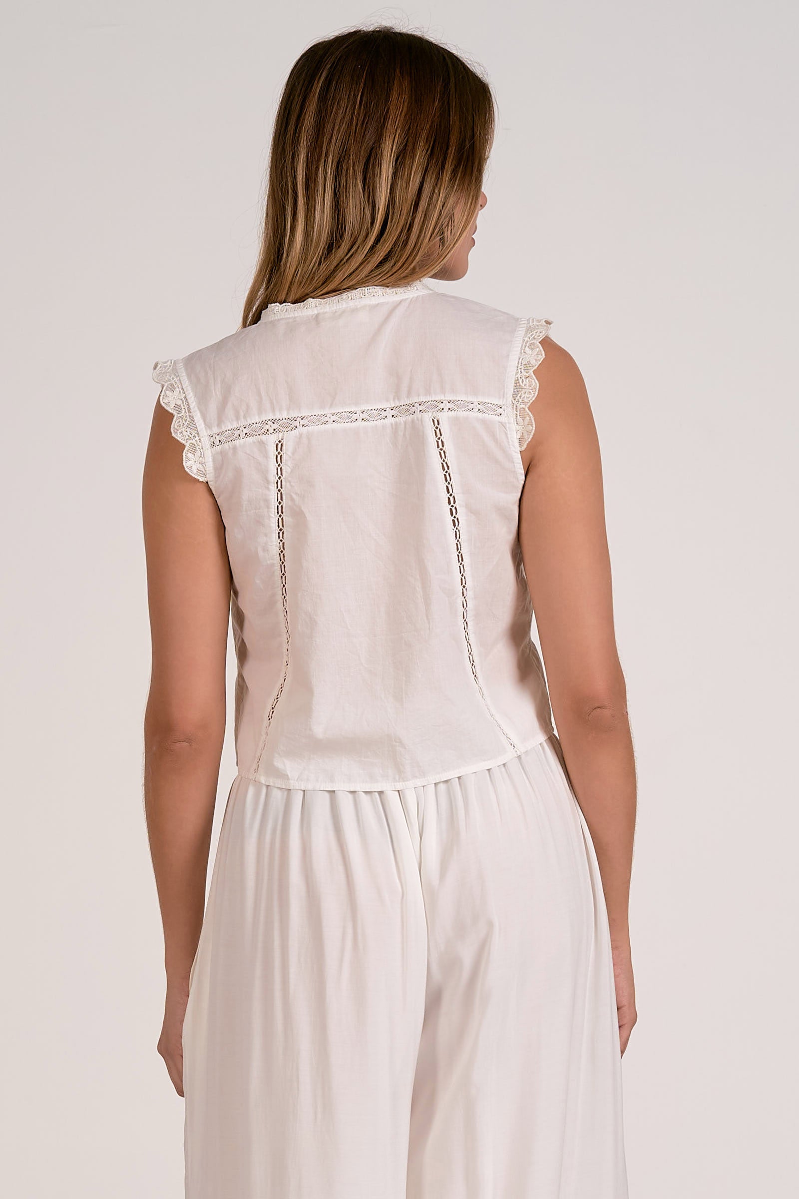 Woman wearing a white sleeveless top and pants with lace details on a plain background