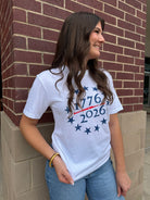 Woman wearing a white t-shirt with '1776 2026' design, standing against a brick wall.