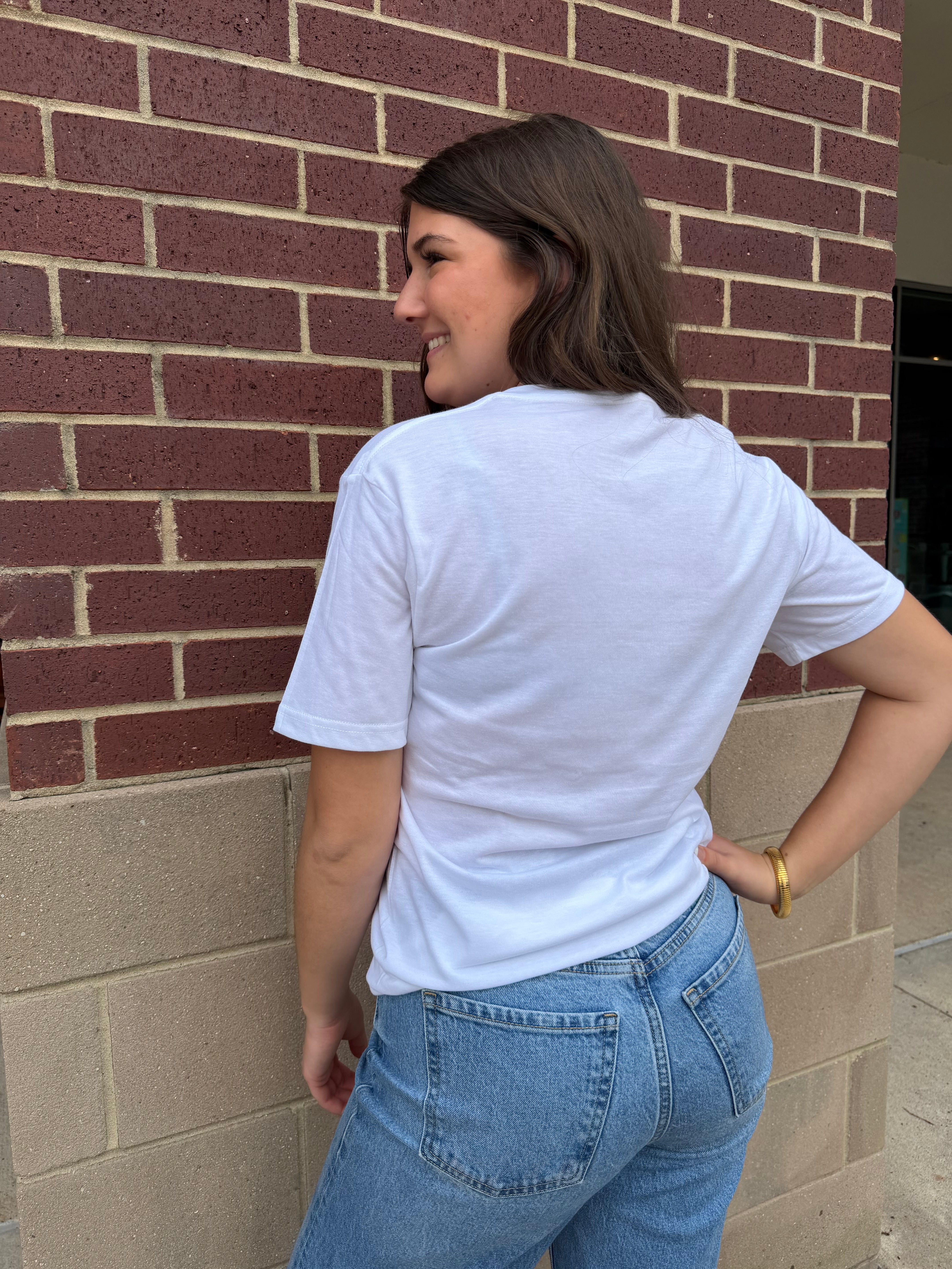 Person wearing a white t-shirt and blue jeans standing against a brick wall.