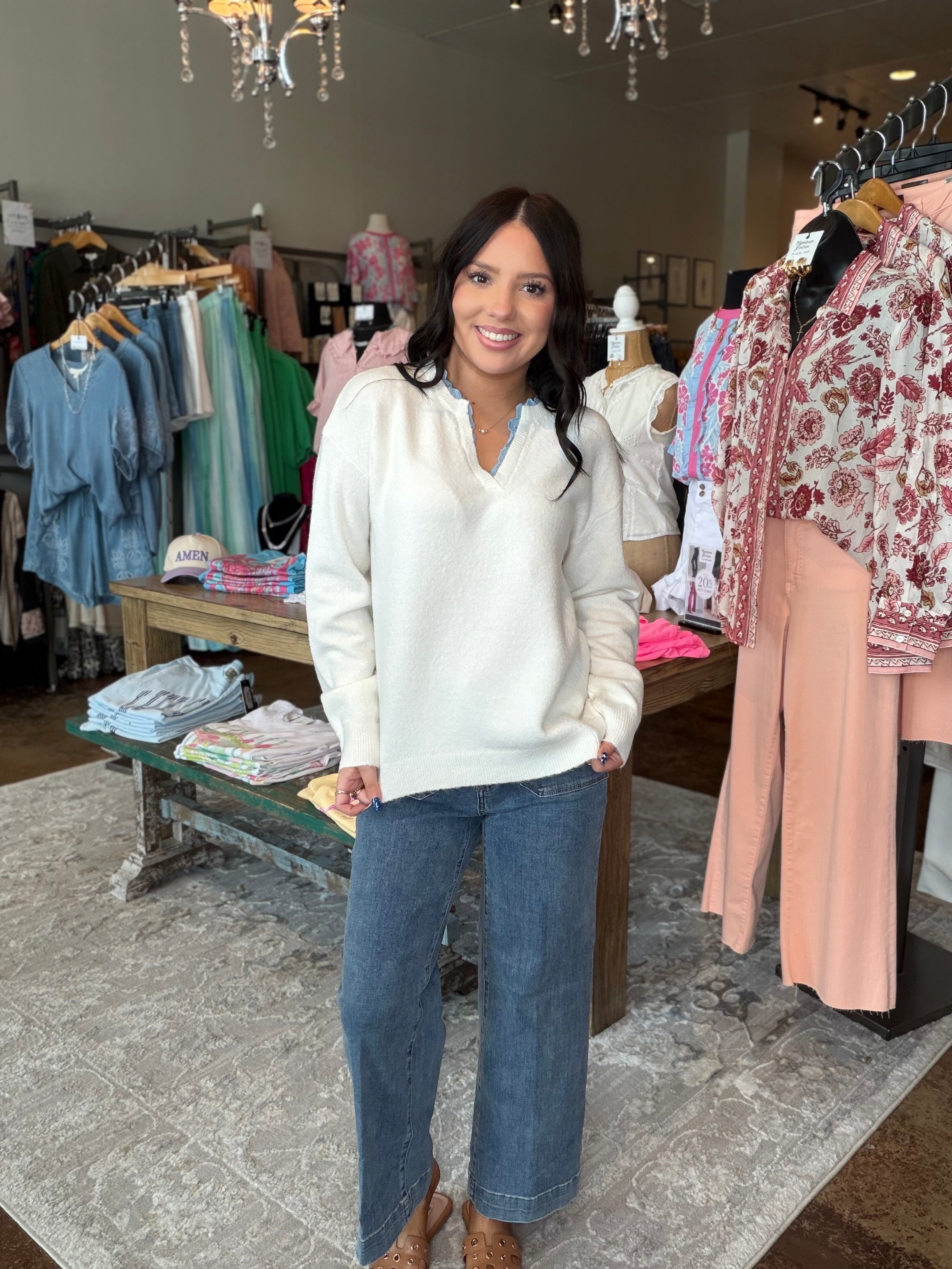 A woman stands in a clothing boutique wearing a cream V-neck knit sweater and wide-leg blue jeans, with racks of apparel and store fixtures visible around her.