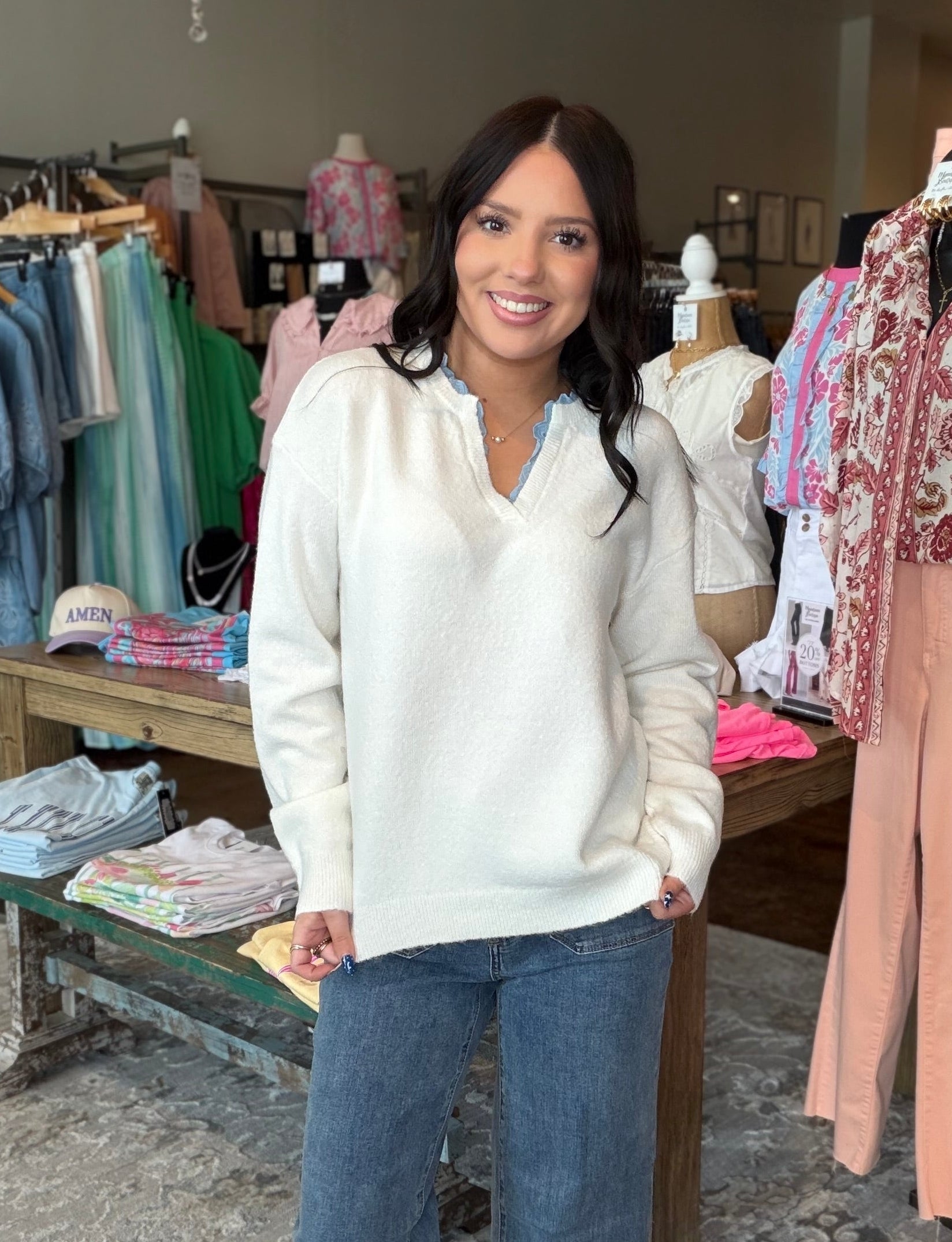 A woman stands in a clothing boutique wearing a cream V-neck knit sweater and wide-leg blue jeans, with racks of apparel and store fixtures visible around her.