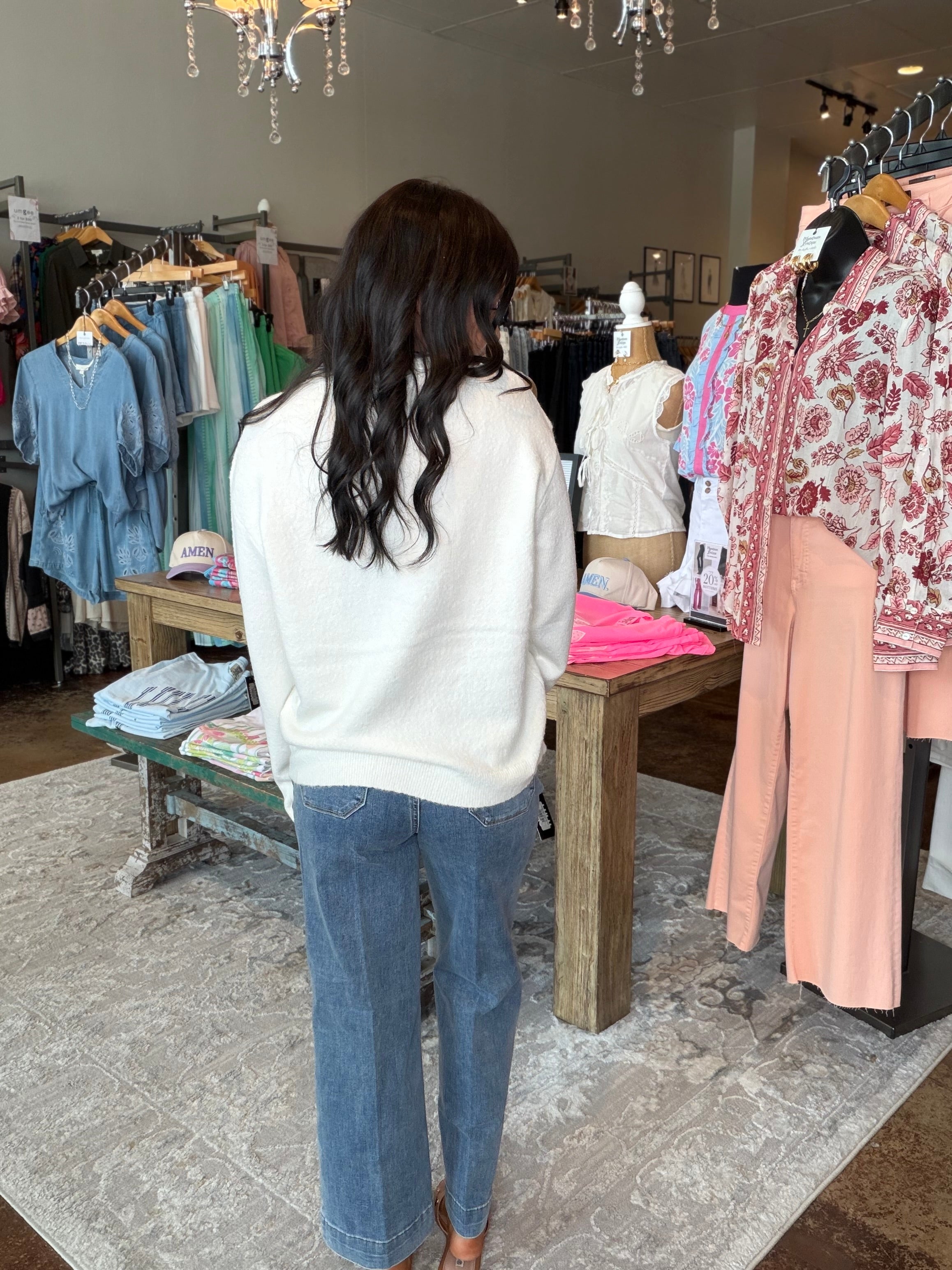 A woman with long dark hair stands with her back to the camera inside a clothing boutique, wearing a cream knit sweater and blue wide-leg jeans. Clothing racks with various garments are visible on both sides of the store.