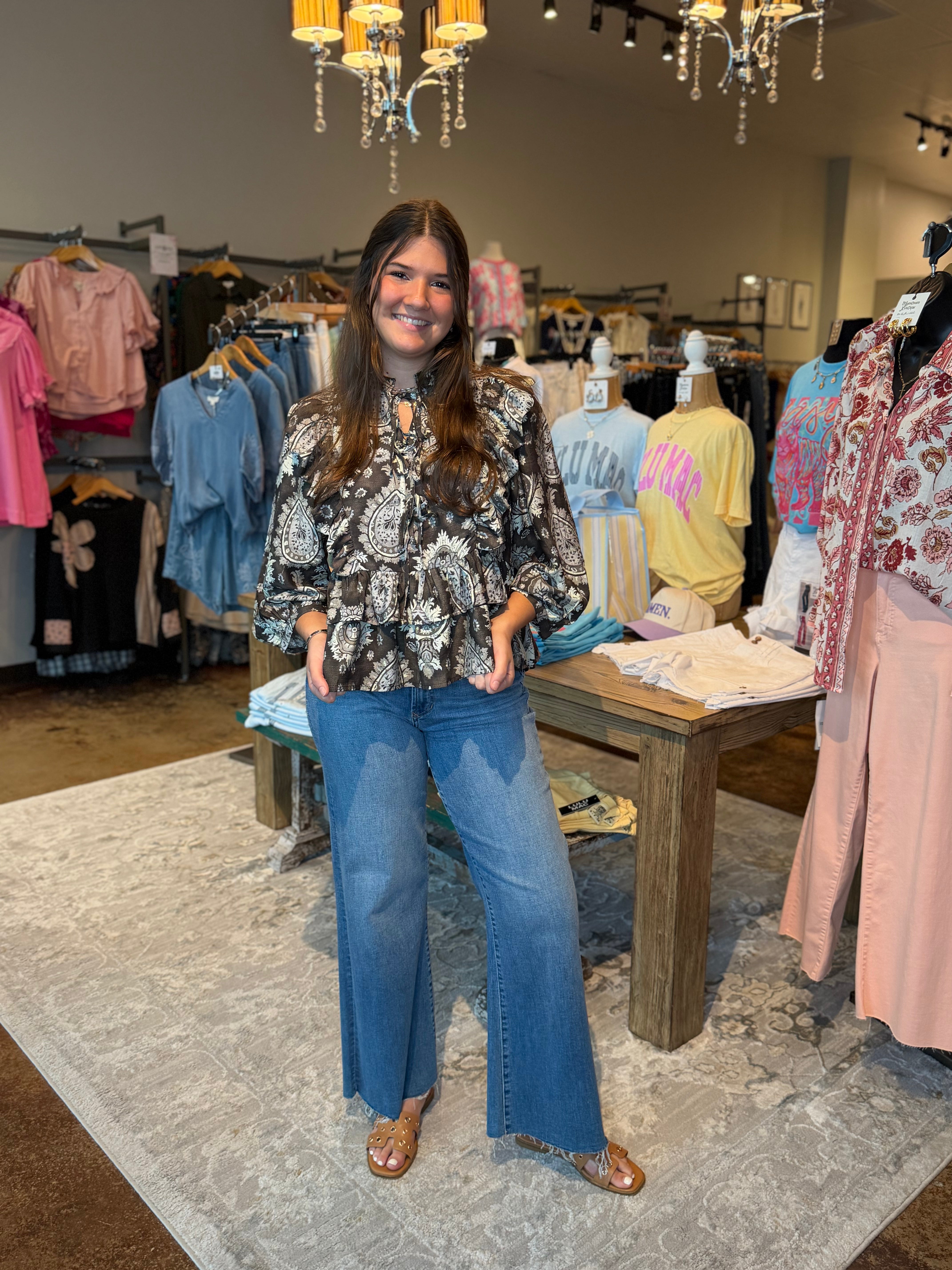 A woman stands in a clothing boutique wearing a black and white paisley patterned long-sleeve blouse with blue wide-leg jeans and brown flat sandals. The store has clothing racks, mannequins, a wooden table, and hanging chandeliers in the background.
