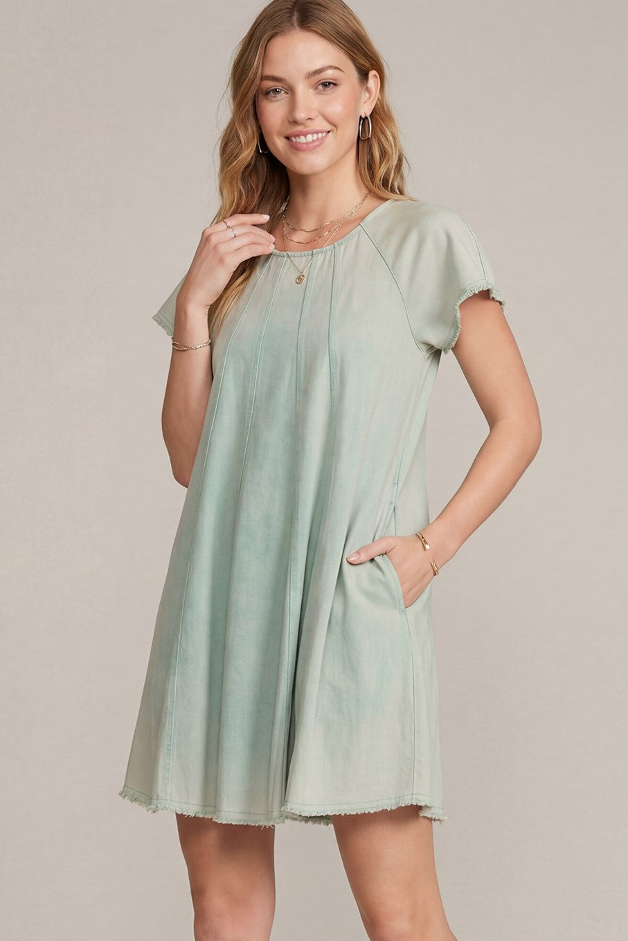 A woman modeling a sage green short-sleeve shift dress with a relaxed fit, side pockets, and raw-edge hems against a solid beige studio background.
