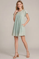 A woman modeling a light sage green short-sleeve shift dress with side pockets, paired with tan heeled sandals, against a solid beige studio background.