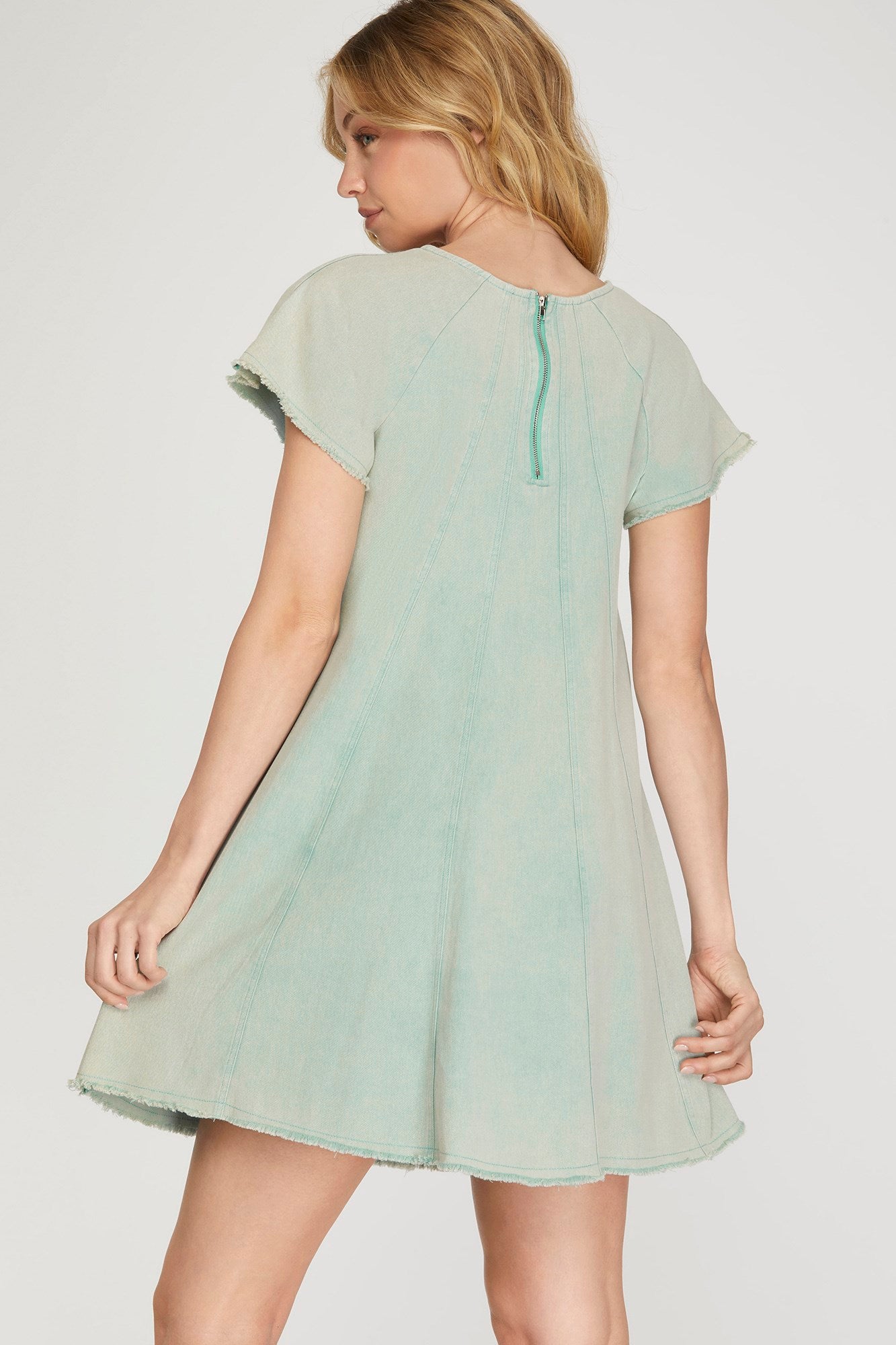 A woman wearing a light sage green short-sleeve dress, shown from the back against a plain light gray studio background.