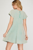 A woman wearing a light sage green short-sleeve dress, shown from the back against a plain light gray studio background.