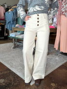 A person wearing off-white wide-leg pants with a high waist and five front buttons, paired with a gray patterned sweater and brown shoes, standing in a clothing store.