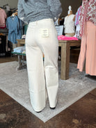 A person standing in a clothing store wearing off-white wide-leg pants and a gray top, shown from the back. The pants have a visible size tag on the back waistband and are photographed in a retail interior with racks, a table, and a rug around them.