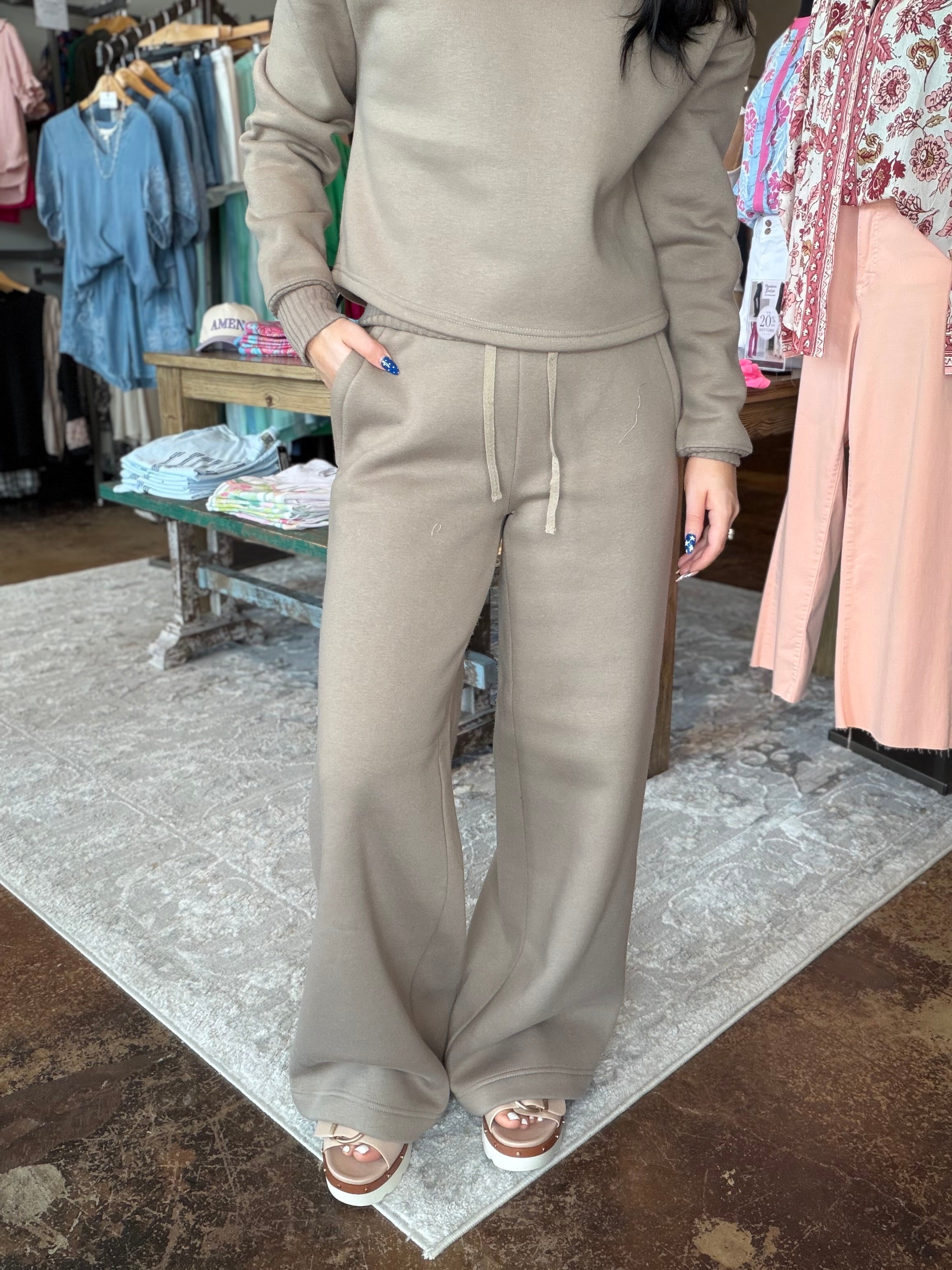 A person wearing a matching taupe-beige lounge set consisting of a cropped long-sleeve top and wide-leg drawstring pants, standing in a clothing boutique.