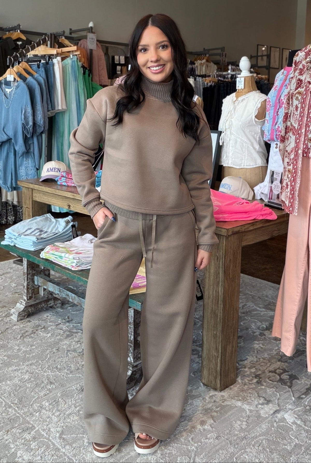 A woman modeling a matching taupe brown lounge set consisting of a turtleneck top and wide-leg sweatpants, standing inside a clothing boutique.