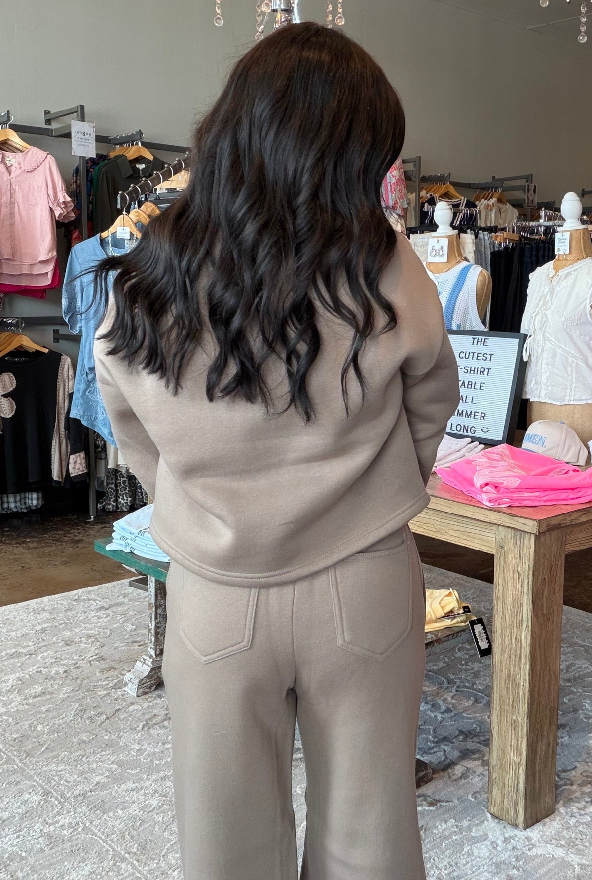 A woman with long dark wavy hair stands in a clothing boutique wearing a beige knit matching set consisting of a cropped sweatshirt and wide-leg pants. She is viewed from behind, with racks of colorful clothing, mannequins, and hanging lamps visible in the background.