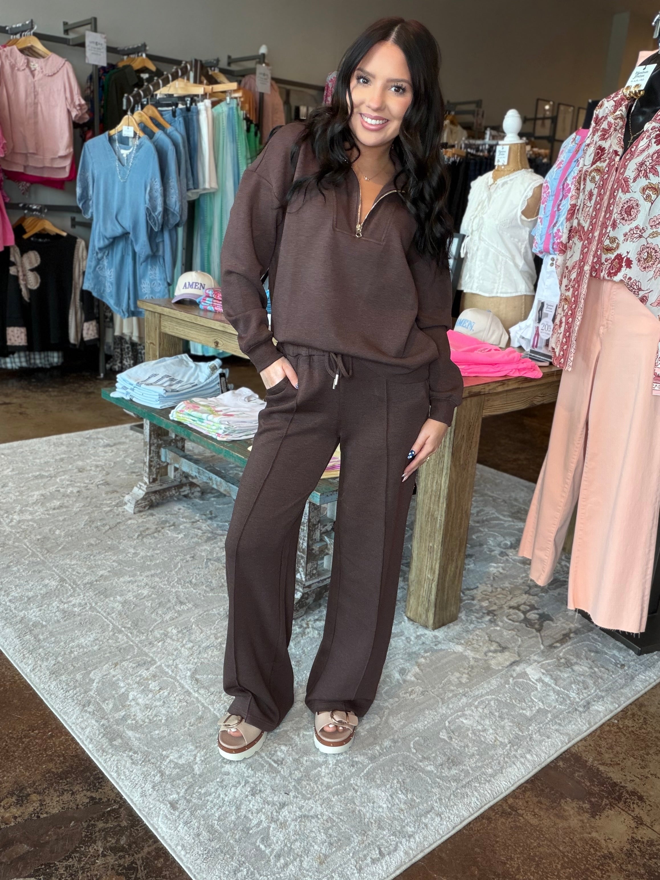 A woman modeling a matching brown lounge set consisting of a half-zip long-sleeve top and wide-leg sweatpants, standing inside a clothing boutique.