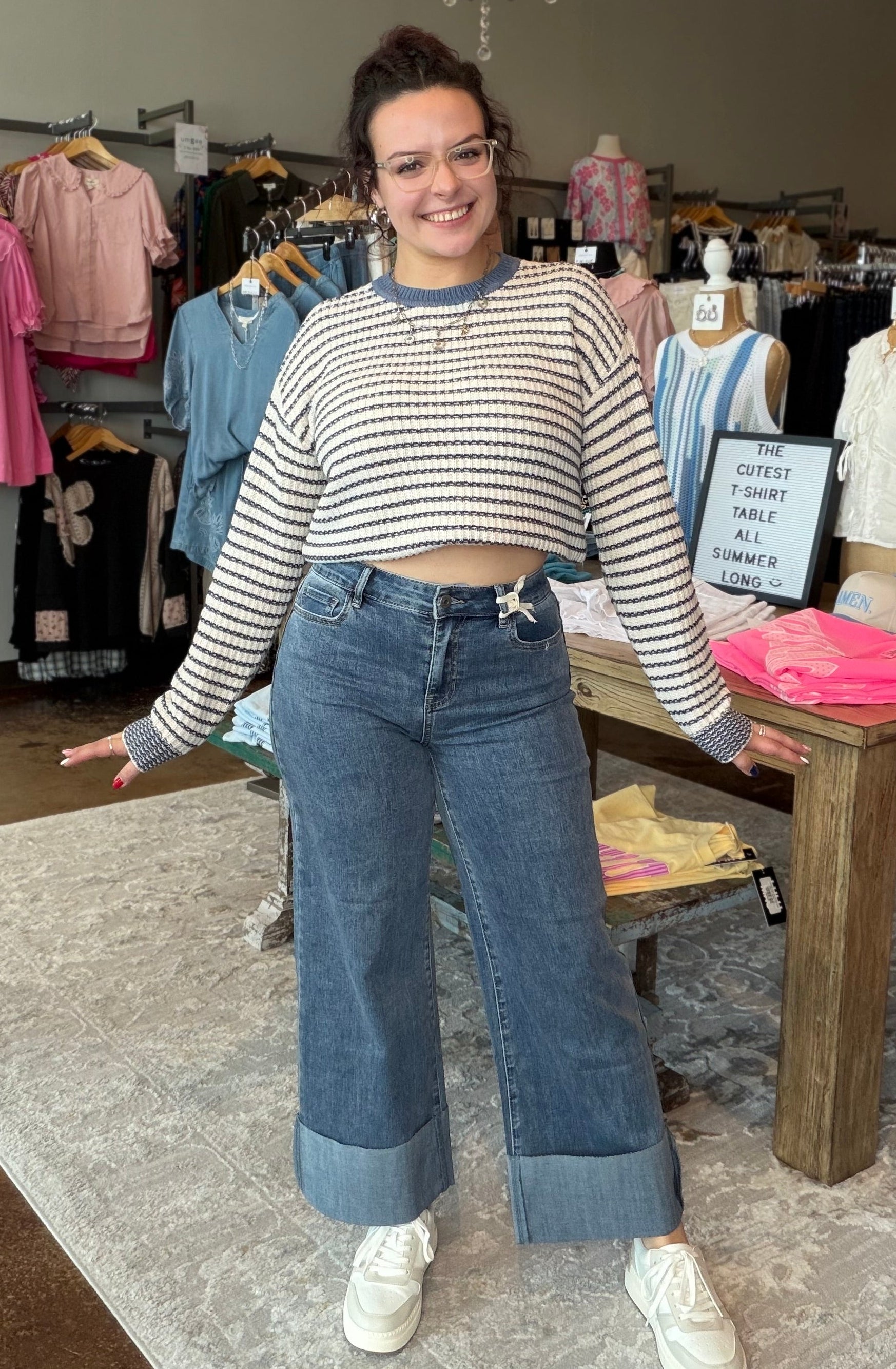A woman modeling a striped long-sleeve crop top and wide-leg blue jeans with rolled cuffs, standing inside a clothing boutique.