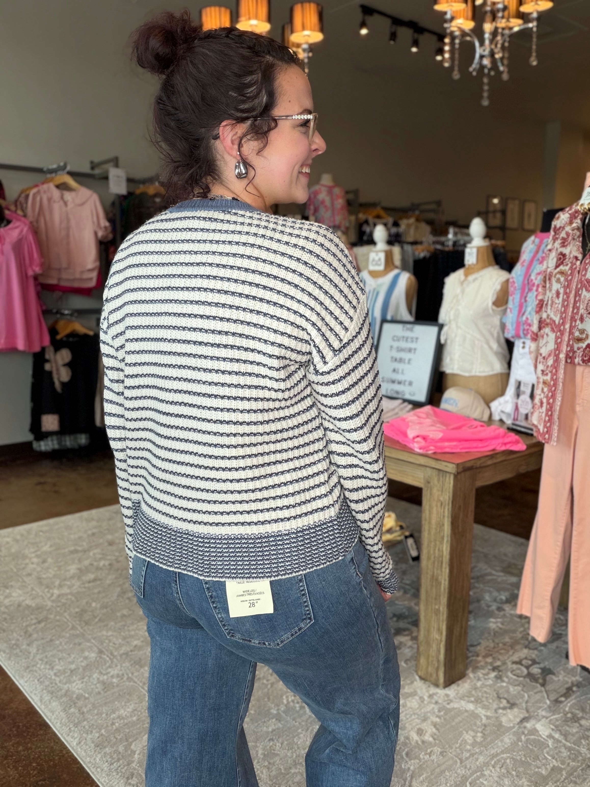A woman wearing a cream and navy blue horizontally striped knit sweater and blue jeans, standing inside a clothing boutique.